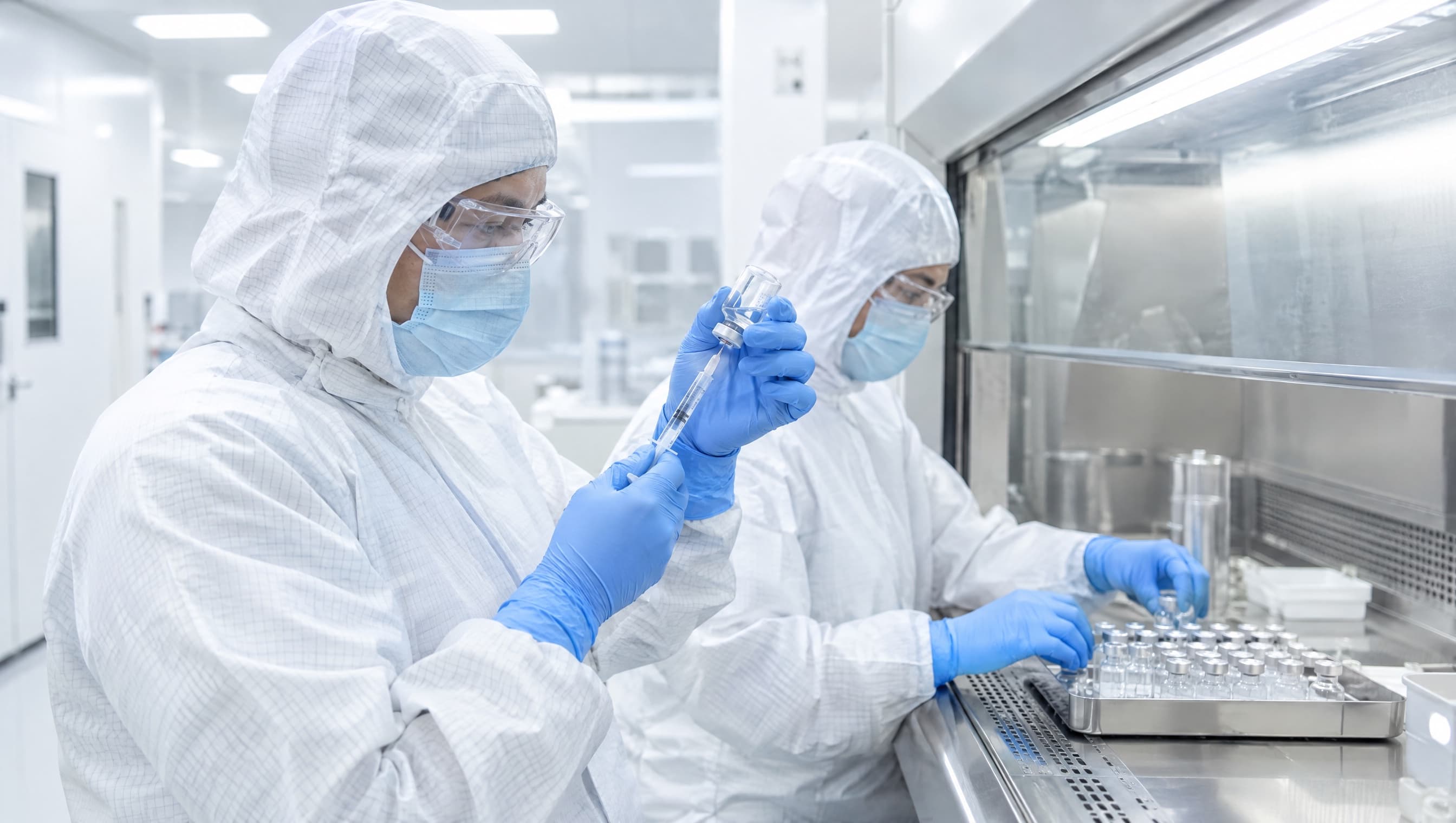 Two technicians in cleanroom gowns and gloves preparing vials at a compounding hood